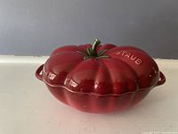 Photo of closed red ceramic bowl with tomato-shaped lid, green stem handle, Staub branding visible on top.