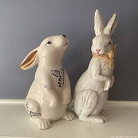 Two decorative rabbit figurines, one ceramic with black leaf designs and glossy finish, one textured white resin-like material with yellow neck ribbon.