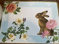Close-up of a full-size placemat featuring a brown rabbit surrounded by flowers on a light blue background with cork backing visible on the edges.