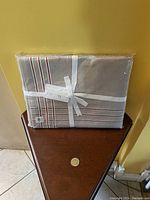 William Sonoma tablecloth in original clear plastic packaging tied with white ribbon, displayed on a small wooden side table. Shows beige fabric with red and white plaid stripes.
