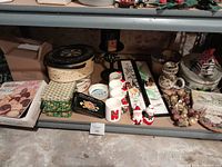 Bottom basement shelf with variety of decor items, tins, mugs, and candle box