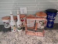 Photo showing three ceramic cannisters with wooden lids decorated with botanical designs, two tall ceramic oil and vinegar bottles with cork stoppers, vintage Tetley tea tin, and bamboo and metal napkin holder arranged on kitchen counter.