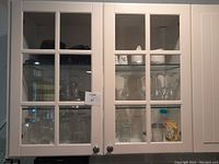 Closed cabinet showing contents behind glass doors including various drinking glasses, wine glasses, white ceramic pieces, and some colored glassware