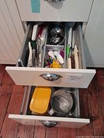 Photo showing two white kitchen drawers pulled out with various kitchen utensils and measuring tools inside, including colanders, spatulas, and whisks.