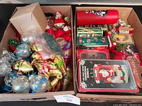 Two boxes containing a variety of Christmas ornaments and holiday decorations, including glass balls, wooden ornaments, and a boxed Santa music decorative item.