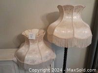 Two off-white vintage Victorian style lamp shades with fringe and floral pattern details, one mounted on a lamp stand.