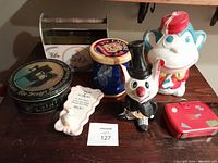 View of the full lot showing plastic monkey bank, ceramic cat bank, Mary Engelbreit tin, Singer tin, Empire tin, sewing kit, and ceramic scissor holder with duck