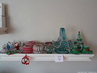 Wide view showing the collection of vintage beaded and safety pin crafted items on a white shelf. Includes angels, candle holders, and Christmas tree shapes.