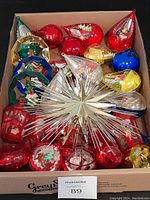 Box containing multiple Jewel Brite vintage Christmas ornaments in various shapes and colors, including teardrops, jewels, wreaths, and diorama lanterns with festive scenes. A large silver and gold aluminum starburst tree topper with spiked design is atop the ornaments.