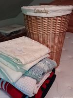 Photo showing tall wicker laundry basket with white fabric liner labeled 'Laundry' beside a stack of assorted blankets and sheer curtains folded underneath.