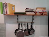 Wood wall shelf with iron brackets, pans hanging beneath, and cookbooks on top