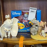 Shelf display with Lenox Dumbo and Timothy Mouse porcelain figurine, boxed Disney Flying Dumbo figure, and an additional Disney Dumbo ceramic figurine with pink-clad elephant and mouse on a yellow base.