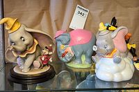Front display of all three items: Giuseppe Armani Dumbo and Timothy Mouse figurine on wooden base, pastel-colored Dumbo coin bank, and Flying Dumbo teapot with crows on cloud base.