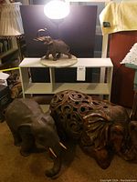 Two large wood carved elephant statues in front, one with damaged tusk, and one with cut out carved pattern. Elephant lamp with marble base above on shelf.