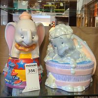 Two Dumbo-themed ceramic cookie jars displayed on glass shelf, showing front view of both jars and condition including noted chip on Timothy Mouse figure.