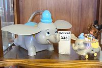 Front view of the large vintage Disney Dumbo popcorn holder next to the smaller Dumbo ride character figurine on wheels, displayed on wooden shelf.
