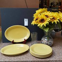 Full lot showing two yellow Fiesta serving platters, four yellow Fiesta dinner plates, one 7" tall clear glass vase, one 10" tall hourglass clear glass vase with faux sunflowers and decorative stones