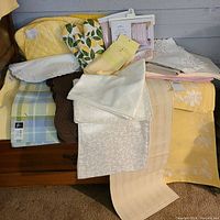 Full lot image showing assorted table linens including placemats, napkins, runners in yellow, cream, white, plaid, and floral patterns.
