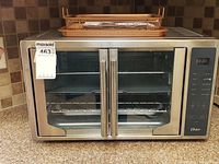 Full view of Oster stainless steel air fry oven with French double doors, showing the control panel with digital display and buttons. Oven interior visible with wire racks inside.