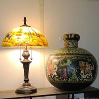 Photo showing the hand-painted urn with animal and people motifs beside the lamp illuminated to reveal the painted wildlife on the glass shade.