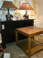 Photo showing wooden side table with brass accents in front, black wood bookcase behind, pair of resin elephant lamps on top of bookcase, and white ceramic elephant plant pot between the lamps.
