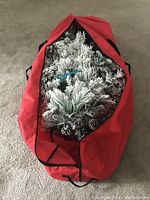 Partially unzipped red wheeled storage bag showing the top portion of the fully flocked artificial Christmas tree with attached pinecones inside.