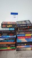 Two stacked piles of paperback and hardback fiction books by various authors including Dean Koontz, Sandra Brown, John Grisham.