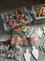 Tote with multi-colored candy bead garlands, bons of tie bags with gingerbread men ornaments, snowflake cookie ornaments, and striped wooden cookie ornaments placed on carpet