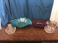 Photo showing blue art glass bowl, purple decor plate, clear crystal pitcher and candy dish displayed on wood surface