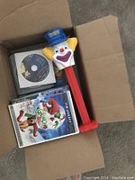 Top view of open cardboard box showing assorted DVDs and oversized PEZ clown dispenser with blue hat, yellow hair and red stem.