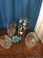 Photo showing five assorted clear and blue decorative glass items on wooden surface against blue and white curtain background.
