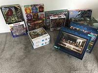 Full view of multiple jigsaw puzzle boxes stacked on a carpet floor, showing a variety of puzzle themes, including birds, music, and classic artworks ranging from 500 to 2000 pieces.
