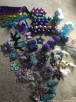 Multiple Mardi Gras themed Christmas decorations including birds, masks, flowers, butterflies, stars, and round ornaments mainly in purple, teal, and gold colors, laid out on floor.