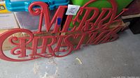Full view of the red metal 'Merry Christmas' sign lying on the floor, showing the ornate script and full wording.