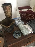 Photo of several metal planters with embossed designs, vase fillers in plastic bags including glass or resin beads of various colors, a red decorative glass or resin bowl planter, and small accessories arranged on a wooden surface.