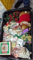 Top down view of large black plastic tote filled with assorted Christmas decorations including floral picks, garlands, packaged ornaments and ribbons, and holiday figurines.