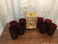 Eight deep red glass tumblers arranged around a decorative glass and brass clock on a wooden surface with patterned curtain backdrop.