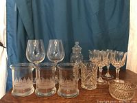 Overview of glassware including ship mugs, stemmed wine glasses, apothecary jar, patterned mug, and small glass bowl arranged on table with blue backdrop.