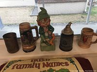 Photo showing five drinking vessels including four steins and one dark drinking cup, plus a ceramic leprechaun figurine playing a flute, all displayed on a windowsill with the Irish Family Names map partially visible beneath.