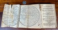 Photo of two open pages showing a double-hemisphere world map printed inside the 1819 geography book. Pages are yellowed, edges worn with a visible tear and crease at the fold. A quarter coin is placed for scale.
