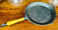 Cast-iron grill pan with round shape and wooden handle, showing the pan lying on a wooden surface.