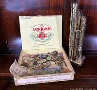 Cigar box open displaying coins, badges, medallions and banknotes alongside the metal coin bank