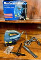 Photo of the Mastercraft orbital jigsaw with its original box in the background, placed on a wooden surface. Also visible are the guide fence, handle attachment, and plastic packet containing blades.