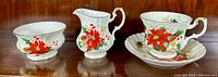 Photo showing all four items: creamer, sugar bowl, cup and saucer with red poinsettia floral design and gold trim.