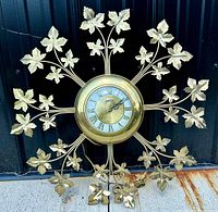 Full front view of vintage brass sunburst wall clock with ivy leaves, black background.