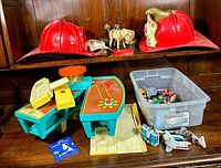 Two red Texaco fire helmets, a windup toy donkey, and a Fisher Price airport playset with various toy cars and Transformers figures arranged in clear bin and on surface below.