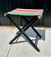 Side view of black painted wood folding camp stool with striped canvas seat showing structure and condition.