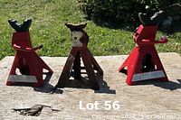 Three jack stands outdoors on a wooden surface, two red painted and one rusted older model, all rated for 3 tons.