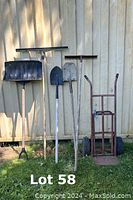 Photo shows six yard tools standing upright against shed wall on grass: black snow shovel, two metal shovels, garden rake, floor squeegee, and two-wheeled dolly.