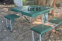 Folded plastic picnic table with attached seats in open, usable position outdoors on ground with wood chip and gravel.